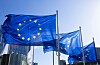 EU-flaggor på flaggstänger framför en modern glasfasad mot blå himmel.