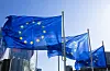 EU-flaggor på flaggstänger framför en modern glasfasad mot blå himmel.