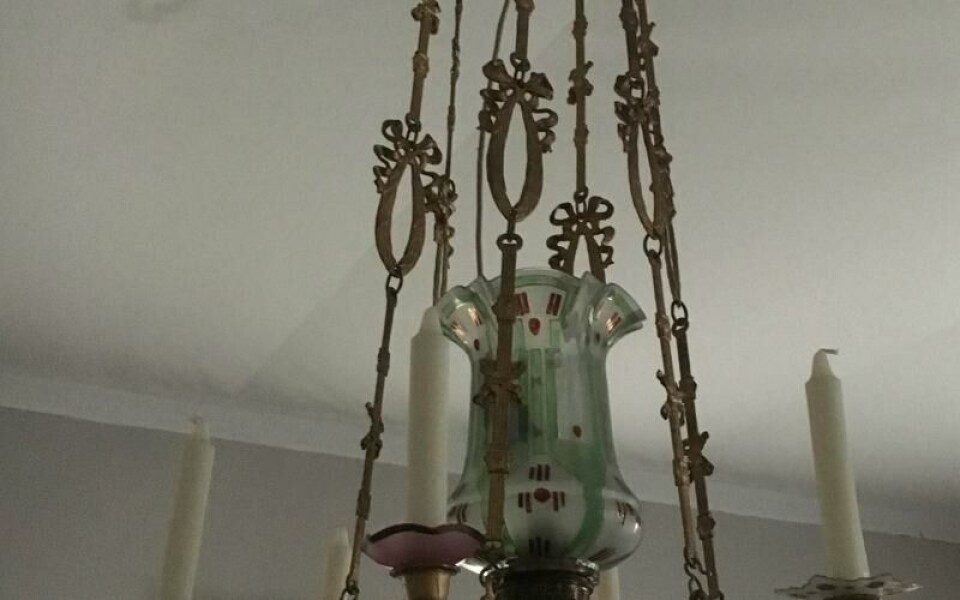 Takkrona med en stomme av bronserad gjuten stomme och majolikatråg kring oljehuset. Stommens ornament har till största delen rötter i empirstilen. Glaskupan har jugenddekor. Takkronan införskaffades till gården Östra Hinders i Önningeby strax efter första världskriget. Då byn fick elektricitet på 1940-talet elektrifierades kronan med en enkel lösning: en glödlampa hängdes ner i lampglaset. Det förekom också att ljushållarna elektrifierades, men här har så inte skett. Kjell EkströmFoto: